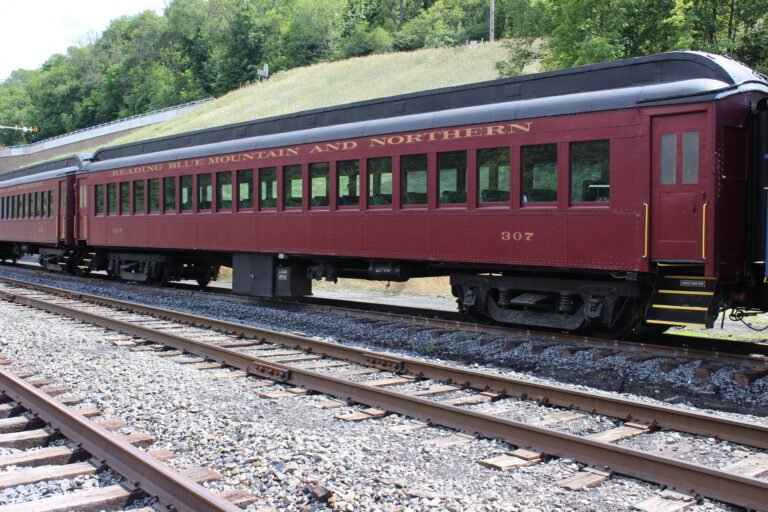Maroon vintage passenger train car labeled 'Reading Blue Mountain and Northern' on railroad tracks with a grassy hill behind it.