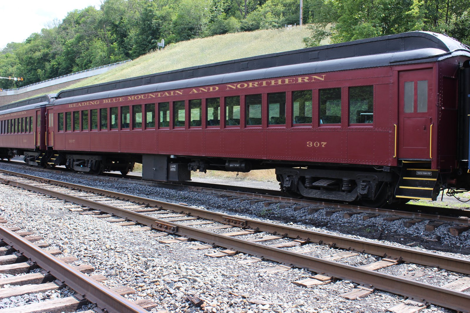 Maroon vintage passenger train car labeled 'Reading Blue Mountain and Northern' on railroad tracks with a grassy hill behind it.