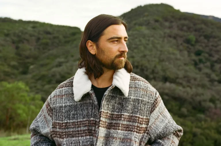 Man with shoulder-length hair and beard wearing a gray plaid coat with a white shearling collar, standing outdoors with hills in the background.