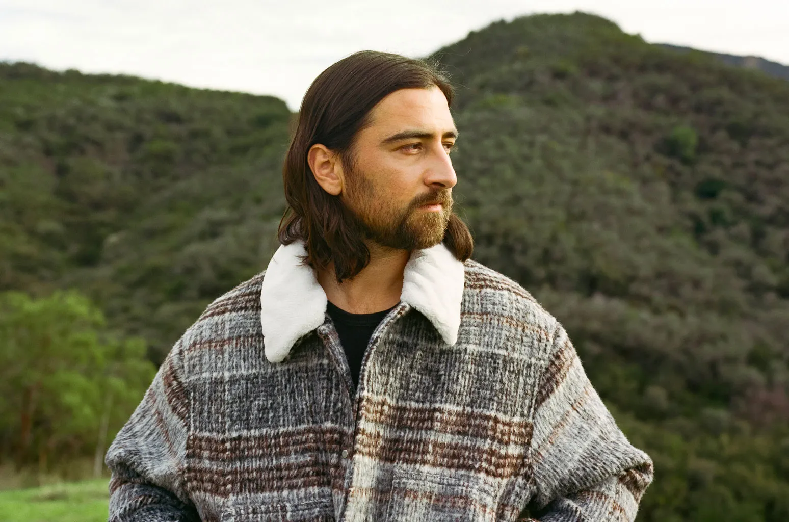 Man with shoulder-length hair and beard wearing a gray plaid coat with a white shearling collar, standing outdoors with hills in the background.